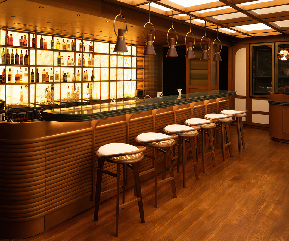A long row of Brummell bar stools with a combination of dark wood, light straw weave (cannage), and a white upholstered seat. The stools are positioned at a green marble-top bar with vertical wood cladding. The atmosphere is cozy and warm, illuminated by a back-lit bottle wall and several bronze suspension lamps at Beefbar Restaurant, Luxembourg.