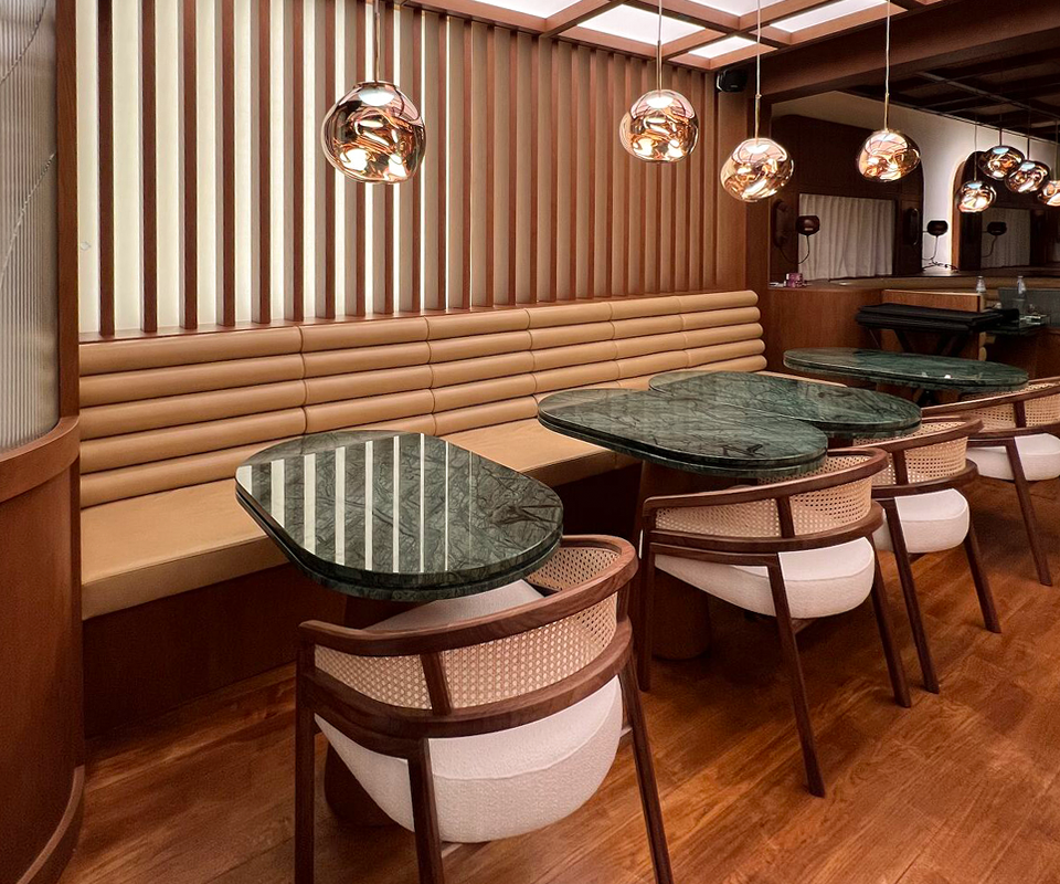 Modern dining area at Beefbar Restaurant featuring dark spencer dining chairs with cane backrests by Wood Tailors Club, green marble tables, a tan leather banquette, and polished metallic pendant lights.