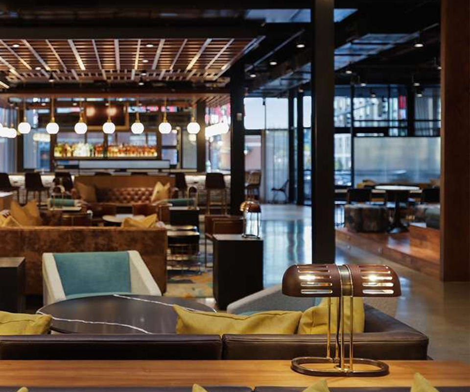 The Turing Table Lamp sitting turned on on a wooden surface in a spacious, modern hotel lounge or restaurant interior at Canopy by Hilton Memphis Downtown.