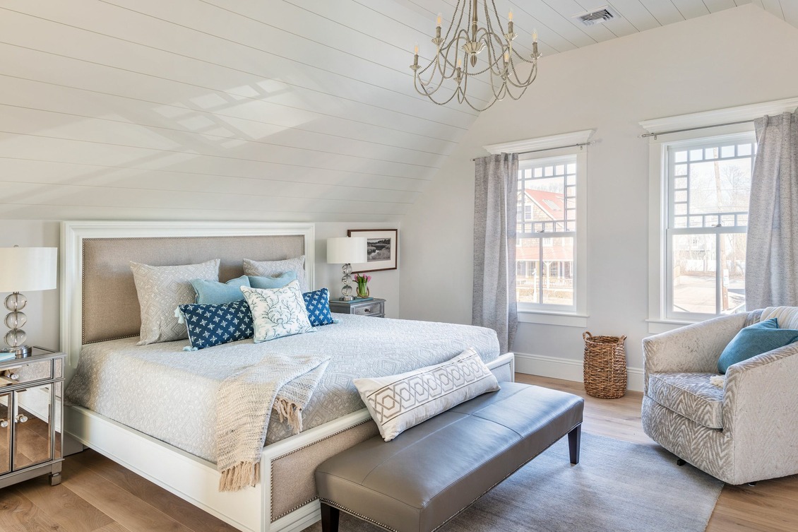 ​A Coastal Grandma Style Home bedroom featuring rattan furniture, including a bed frame and seating, complemented by white linens with subtle patterns, wooden furniture with a distressed finish, and soft lighting from bedside lamps with linen shades.