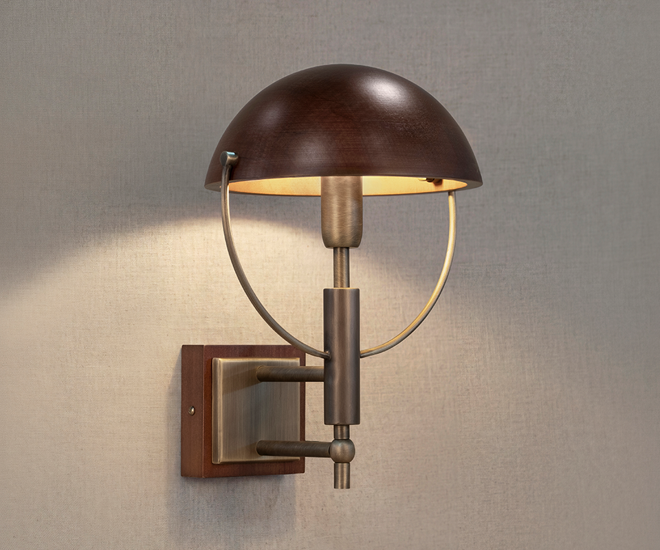 Faraday Wall Lamp by Wood Tailors Club switched on against a beige fabric-textured wall, casting a warm glow through its smoked walnut shade and aged brushed brass structure.