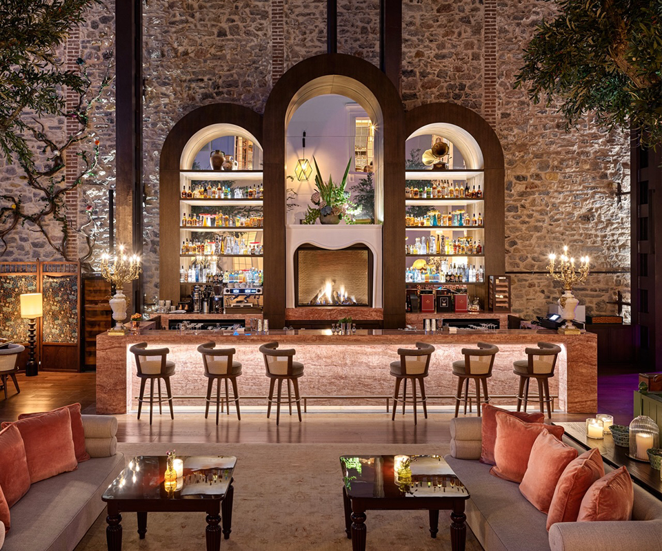 A line of George Bar Chairs by Wood Tailors Club with brown leather upholstery set along an illuminated marble counter inside the rustic-chic Little House bar with stone walls and a cozy fireplace.