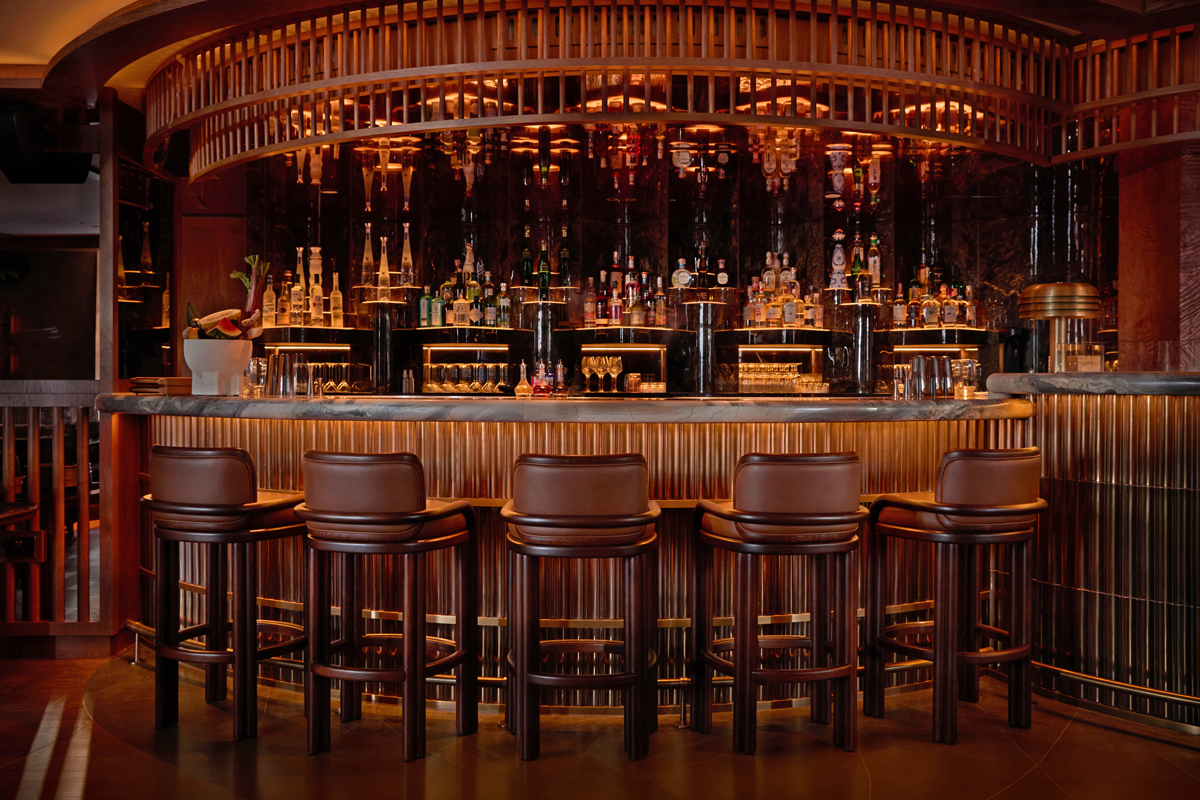Projects Ma na 9 Close-up photograph of the opulent bar counter at MA/NA Mayfair. The circular bar features a veined marble top and a base clad in fluted golden metallic panels that catch the embedded lighting. Lined up in front of the counter are five high bar stools in brown leather with dark wood frames, supplied by Emotional Brands. In the background, the richly illuminated bar shelves display premium spirit bottles against a dark, mirrored backdrop under a curved wooden slat canopy.