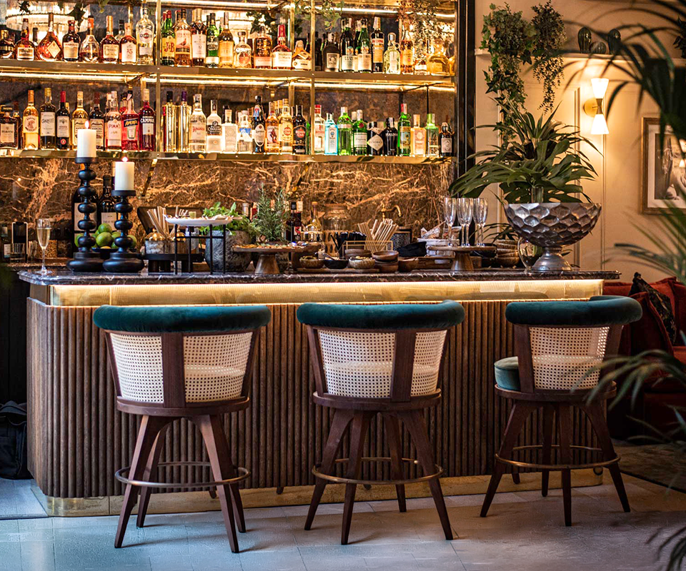 Three George Bar Chairs by Wood Tailors Club featuring walnut wood, woven rattan backrests, and emerald green velvet seats, lined up along a marble bar at Palma Suites Maiorca.
