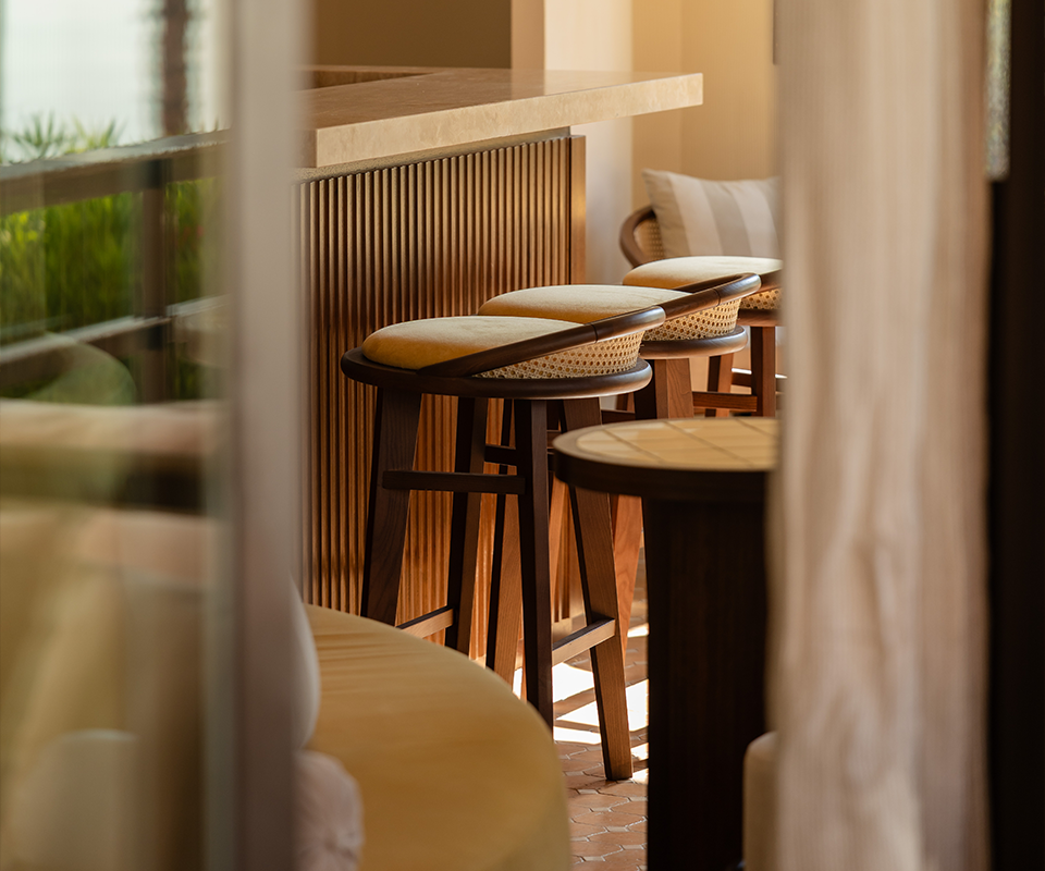 A row of Brummell bar stools with a combination of dark wood, light rattan, and a beige upholstered seat. The stools are lined up at a bar counter made of vertical wood strips. The perspective is slightly restricted, as if through an open door, with natural light coming from the left at Pool House by Studio Quentin, Monte-Carlo, Monaco.