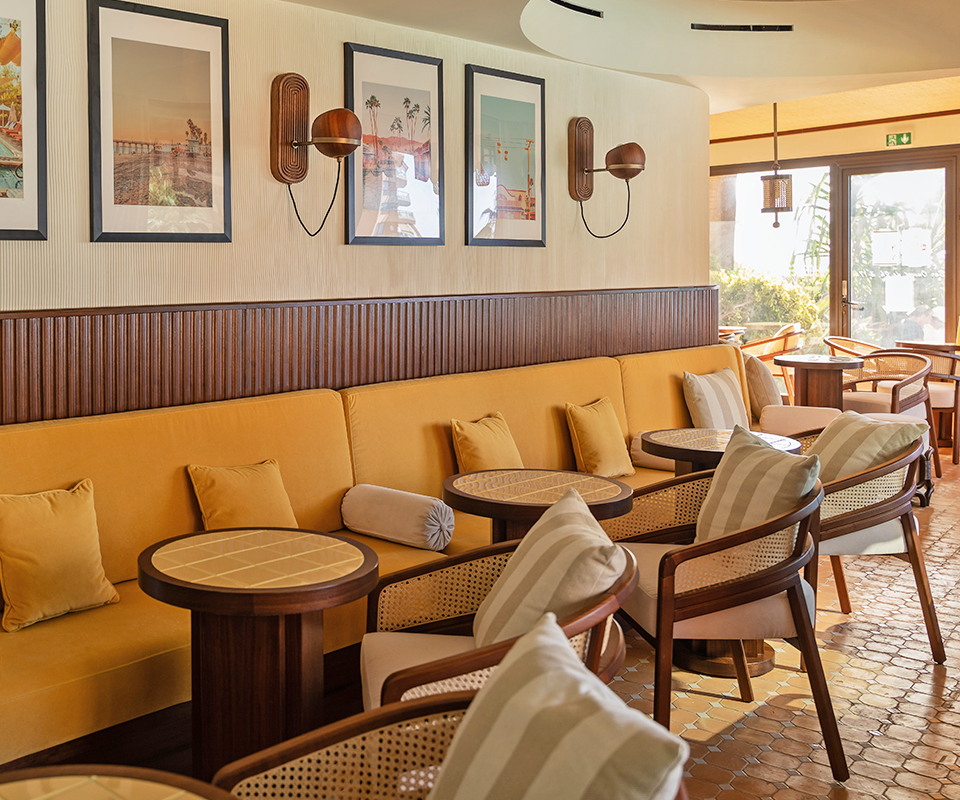 Warm lounge interior for Pool House by Studio Quentin featuring yellow upholstered seating, small round wooden coffee tables, and Wood Tailors Club spencer dining chairs with striped pillows.
