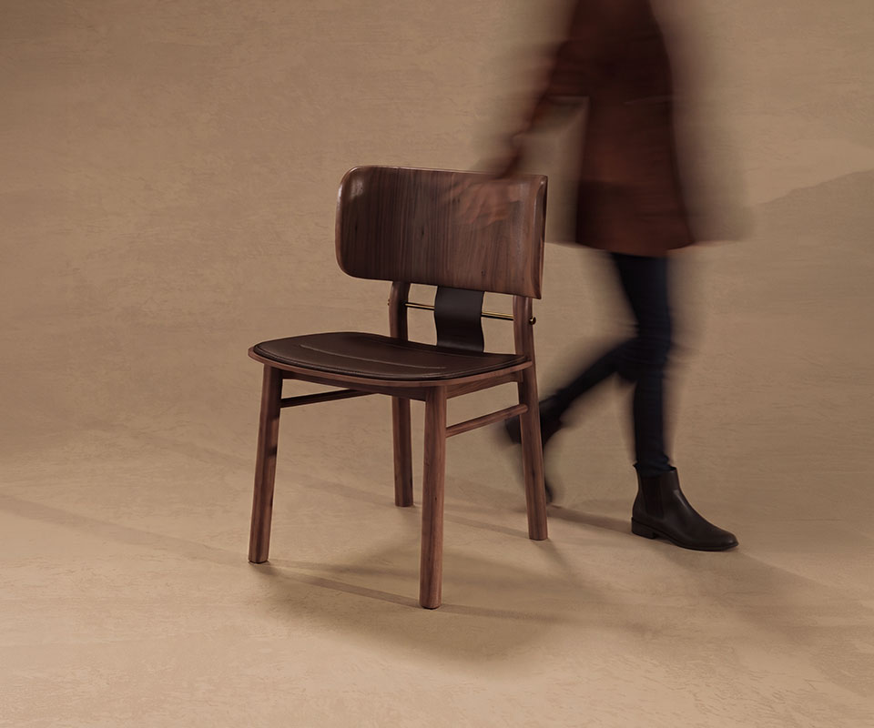 Three-quarter front view of the Arthur Dining Chair in a warm studio setting, with a motion-blurred person walking past in the background.
