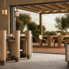 Modern luxury outdoor patio dining area featuring the Benjamin Dining Chair and Portman Dining Table on a wooden deck. Inside, three wooden Benjamin Bar Chairs sit at a stone bar counter under Hamilton Pendant Lamps. A Wodehouse Sideboard decorated with a Hamilton Table Lamp completes the warm, minimalist indoor-outdoor setting.