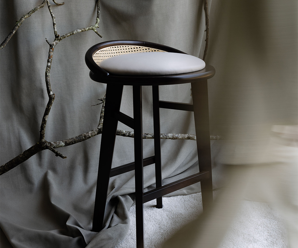 Brummell bar stool in black stained ashwood featuring a light grey upholstered round seat and a curved cane backrest, styled in a minimalist studio setting with neutral fabric backdrop and tree branches.