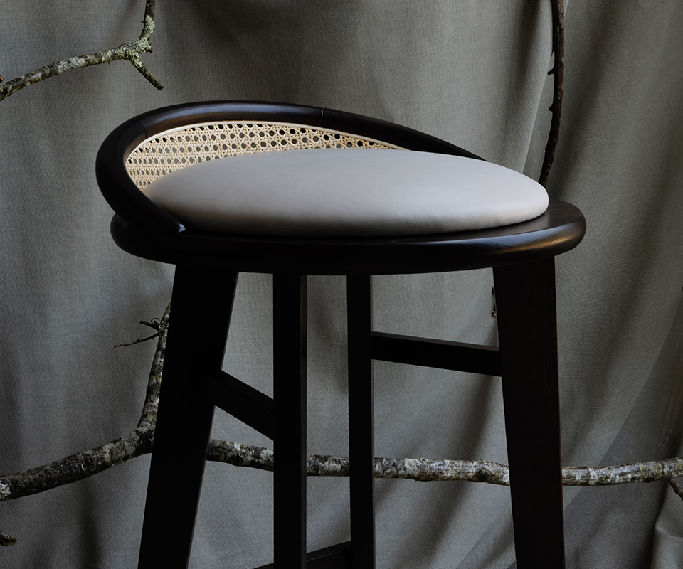 Close-up view of the Brummell bar stool in black stained ashwood, highlighting the light grey cushion seat and woven cane detail on the curved backrest against a draped fabric backdrop.