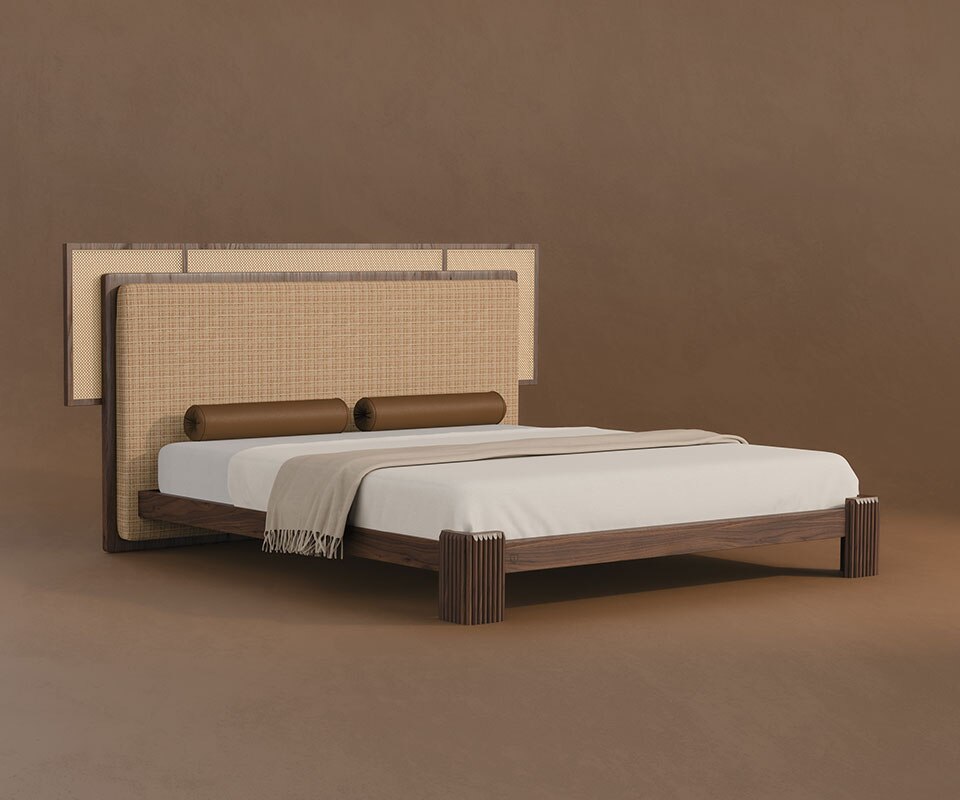 rattan bedroom furniture. Forbes bed in a 45º front view, featuring a fabric and rattan headboard, cylindrical leather bolster pillows, and a walnut wood frame, set against a brown background.