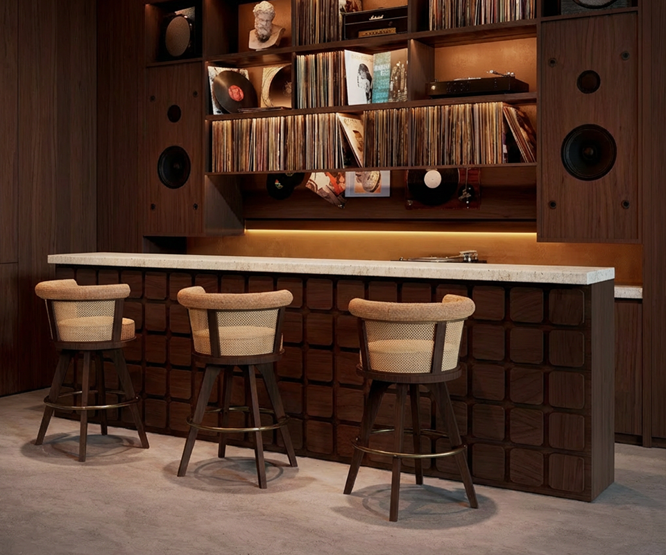 George Bar Chair A side-angle view of three George Bar Chairs positioned along a textured dark wood bar with a white stone countertop. The chairs showcase their mid-century design with tapered walnut legs, brass footrests, and curved rattan backrests. In the background, a large custom wooden shelving unit integrates professional speakers and a vast vinyl record collection under warm, recessed golden lighting. The floor is a smooth, light grey polished concrete, enhancing the modern club atmosphere.
