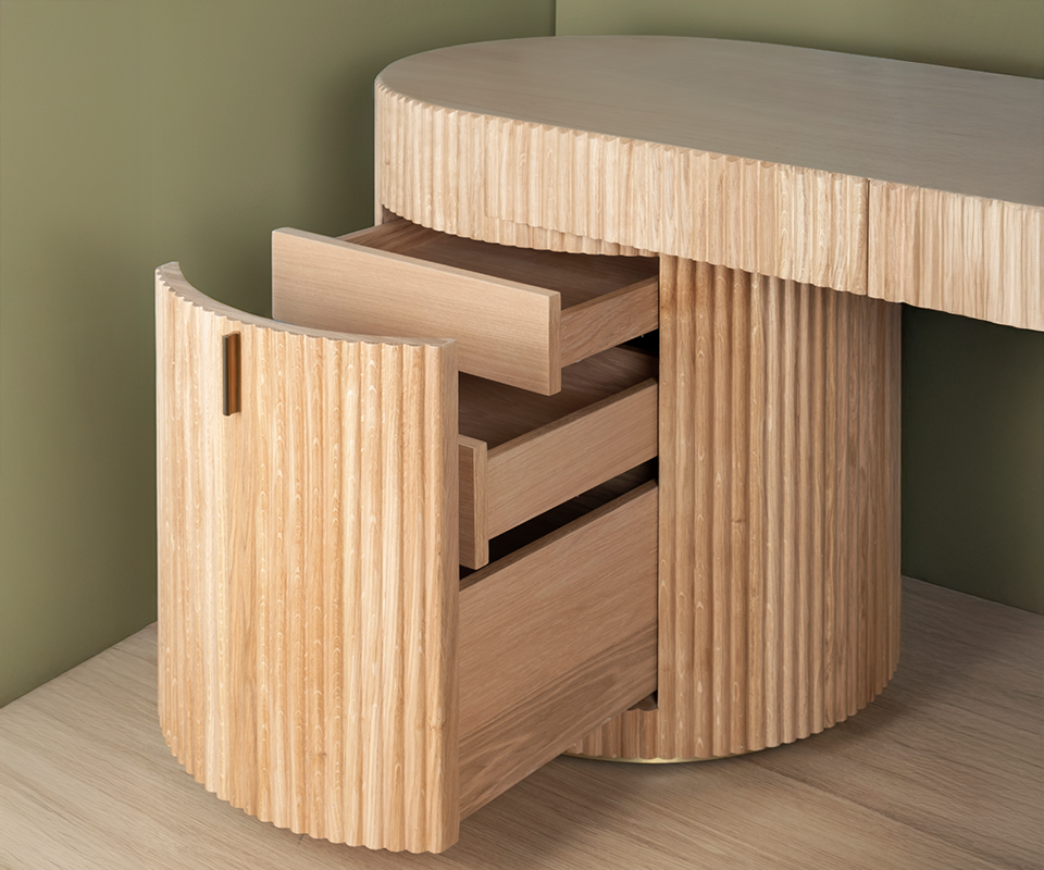 Gifford Desk Close-up of the Gifford desk by Wood Tailors Club in oak. The curved, fluted door of the left cylindrical cabinet is open, revealing three extended inner oak drawers. A small brass pull handle details the door, with the desk resting on a light wooden floor against a soft green wall.