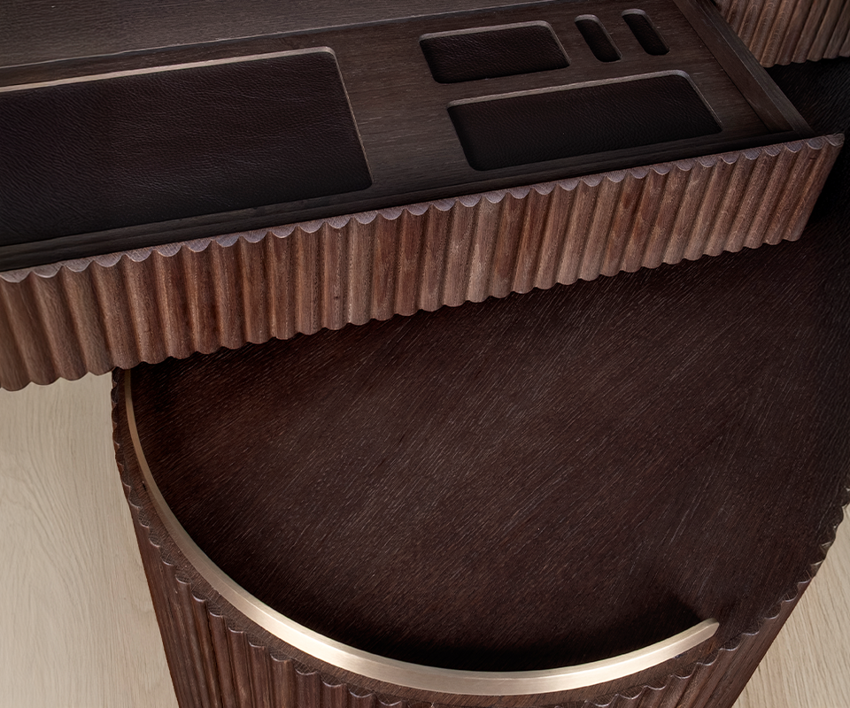 Gifford Desk Detailed close-up of the Gifford desk by Wood Tailors Club in smoked walnut wood. An open drawer displays built-in, dark leather-lined organizational compartments. The image highlights the fluted wood texture of the drawer front and the curved edge of the lower module below, accented with a polished brass trim.
