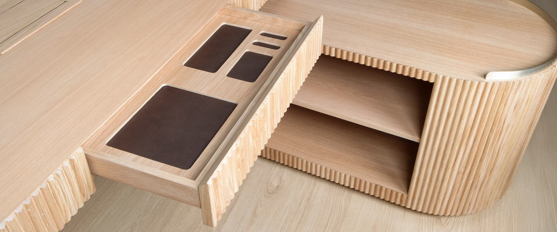 Gifford Desk Close-up detail of the Gifford desk by Wood Tailors Club in oak. The main desktop drawer is pulled open, showcasing dark leather-lined organizer compartments. The fluted texture is prominently visible on the drawer front and on the adjacent lower oak module to the right, which reveals two open storage shelves.