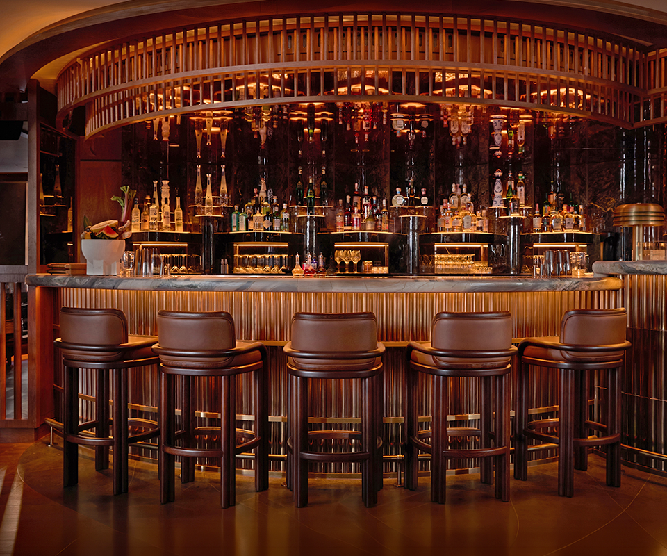 Row of five James Bar Chairs in smoked walnut wood and tan leather lined along a luxury bar counter in a sophisticated, dimly lit restaurant setting.