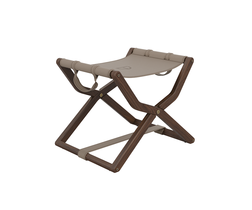 Three-quarter view of the Korda Folding Stool showcasing a walnut wood frame with a rich, deep finish. The design features a beige leather sling seat with elegant strap details and metallic accents, embodying a sophisticated, modern silhouette.