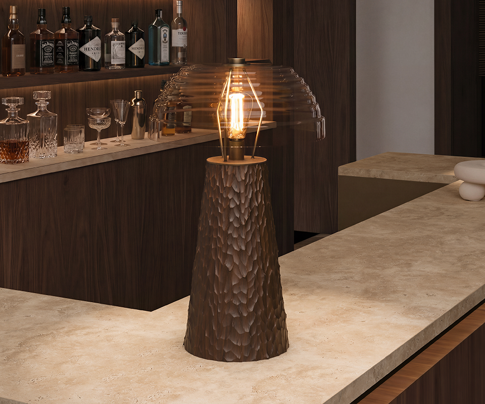 A close-up view of the Lloyd I Table Lamp illuminated on a textured cream marble bar counter, showing the details of its dark hammered wood base and glowing bulb beneath a ribbed extra clear glass shade, with a selection of bottles blurred in the background.