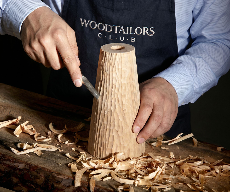 Close-up of a craftsman wearing a Woodtailors Club apron, meticulously hand-carving the textured wooden base of the Lloyd I Table Lamp with a chisel on a workbench surrounded by wood shavings.