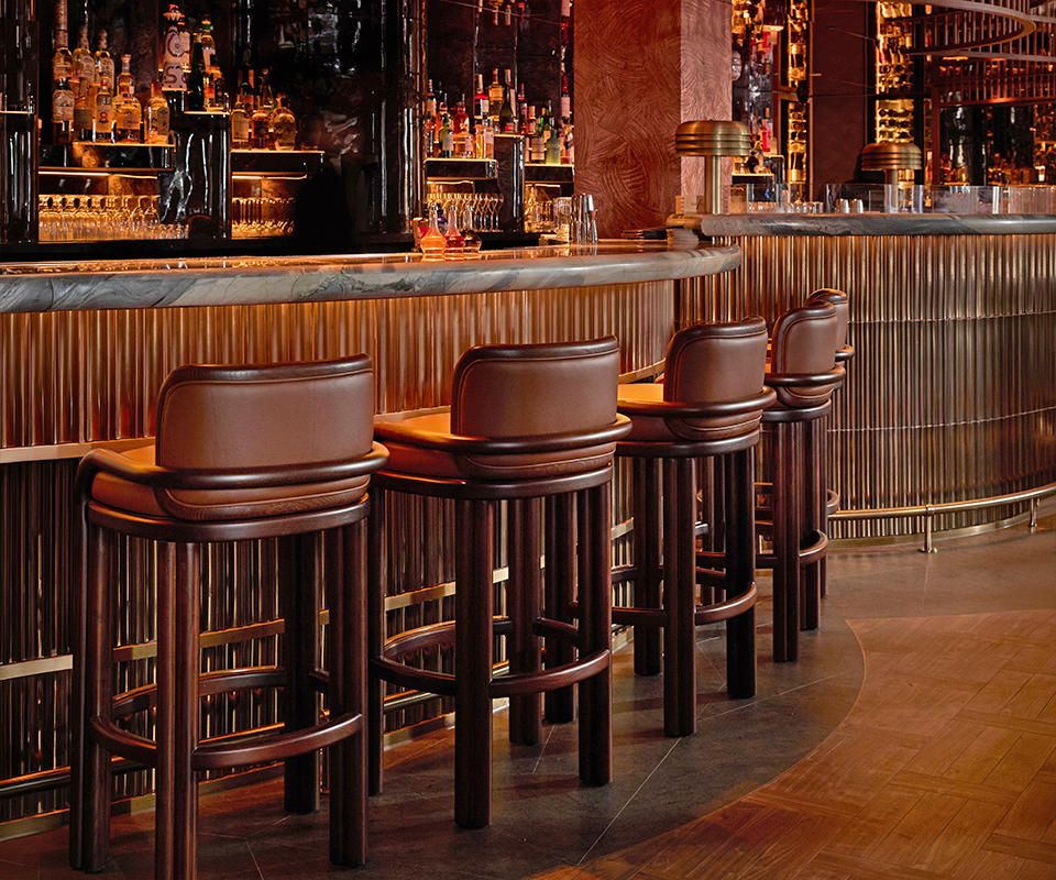 Row of dark wood James Bar Chairs with brown leather upholstery lined up along an elegant, warm-lit marble counter at MA/NA Mayfair, London, UK bar.