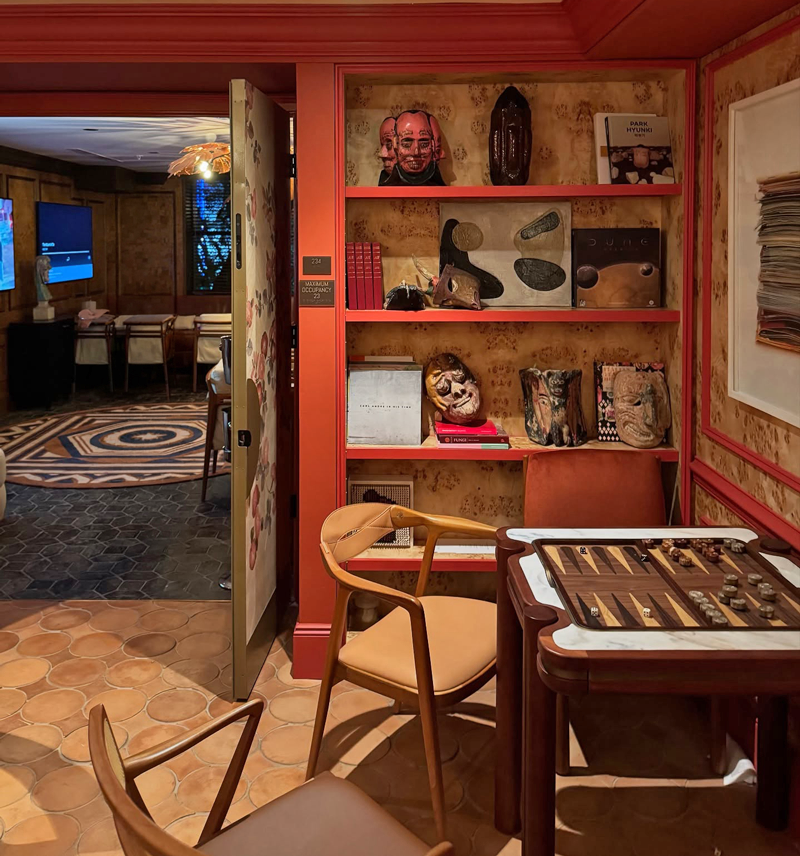 Projects moore hotel 10 A close-up view of a cozy gaming corner inside The Moore Miami Social Club, showcasing a wooden Holland Backgammon Table set up with checkers and dice. Two modern wooden chairs are placed in front of it. In the background, built-in shelves displaying art books, ceramic masks, and sculptures line a burl wood wall with red trim. An open decorated door reveals another room with a geometric rug and a flat-screen TV in the distance.